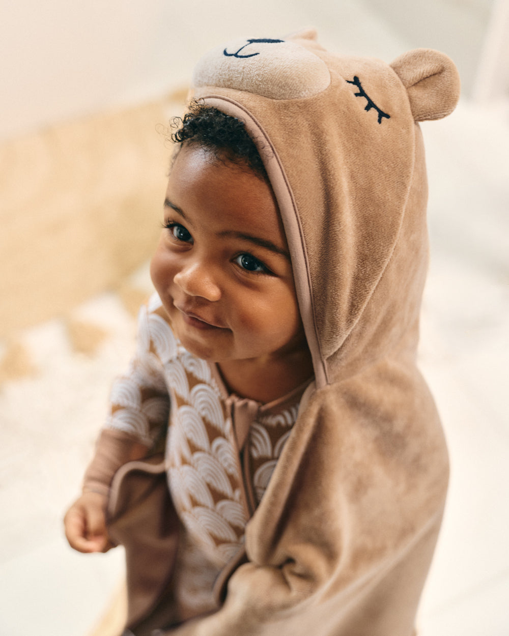 A close-up of a smiling toddler wearing a plush hooded onesie with a bear face design, photographed in a softly lit indoor setting.