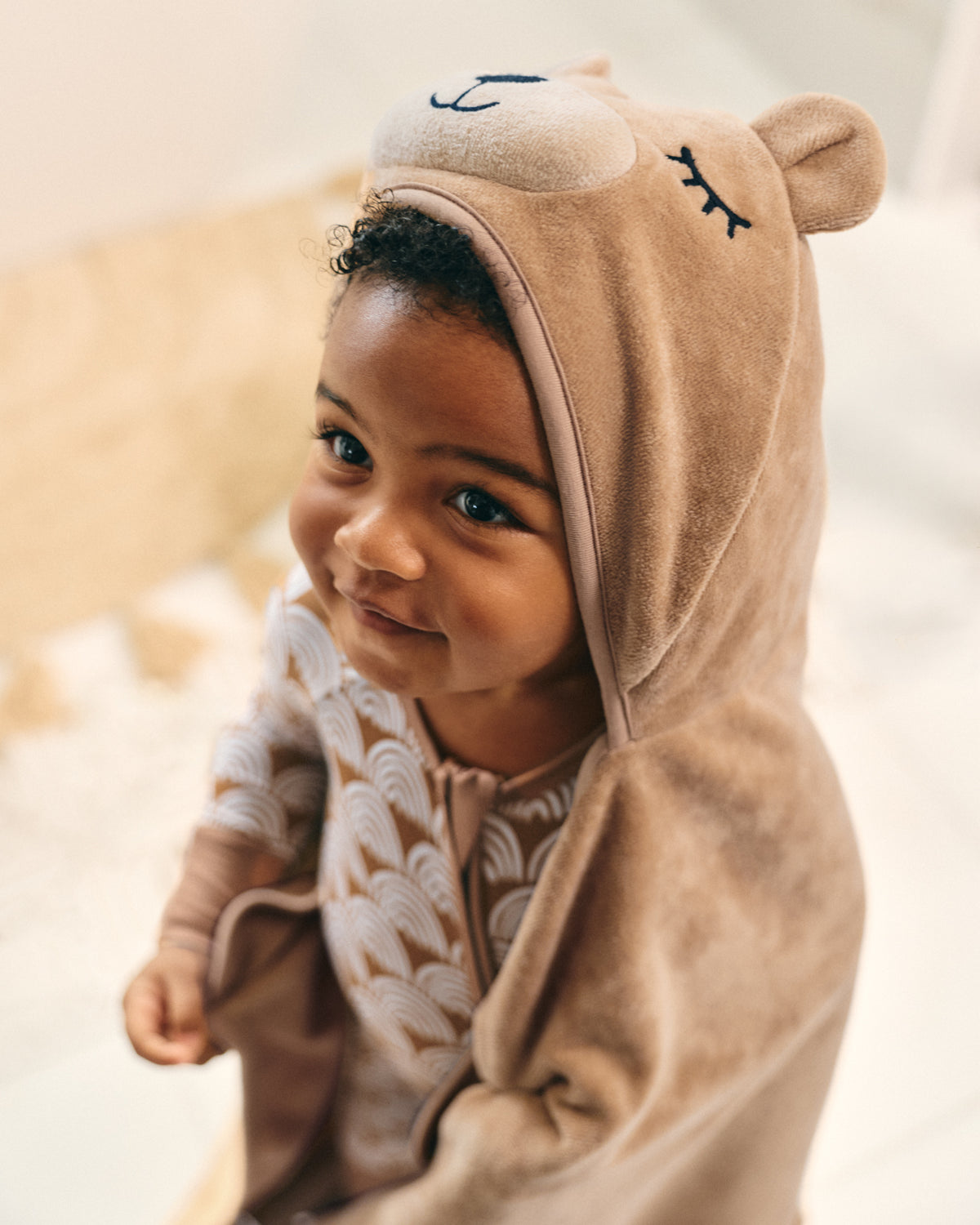 A close-up of a smiling toddler wearing a plush hooded onesie with a bear face design, photographed in a softly lit indoor setting.