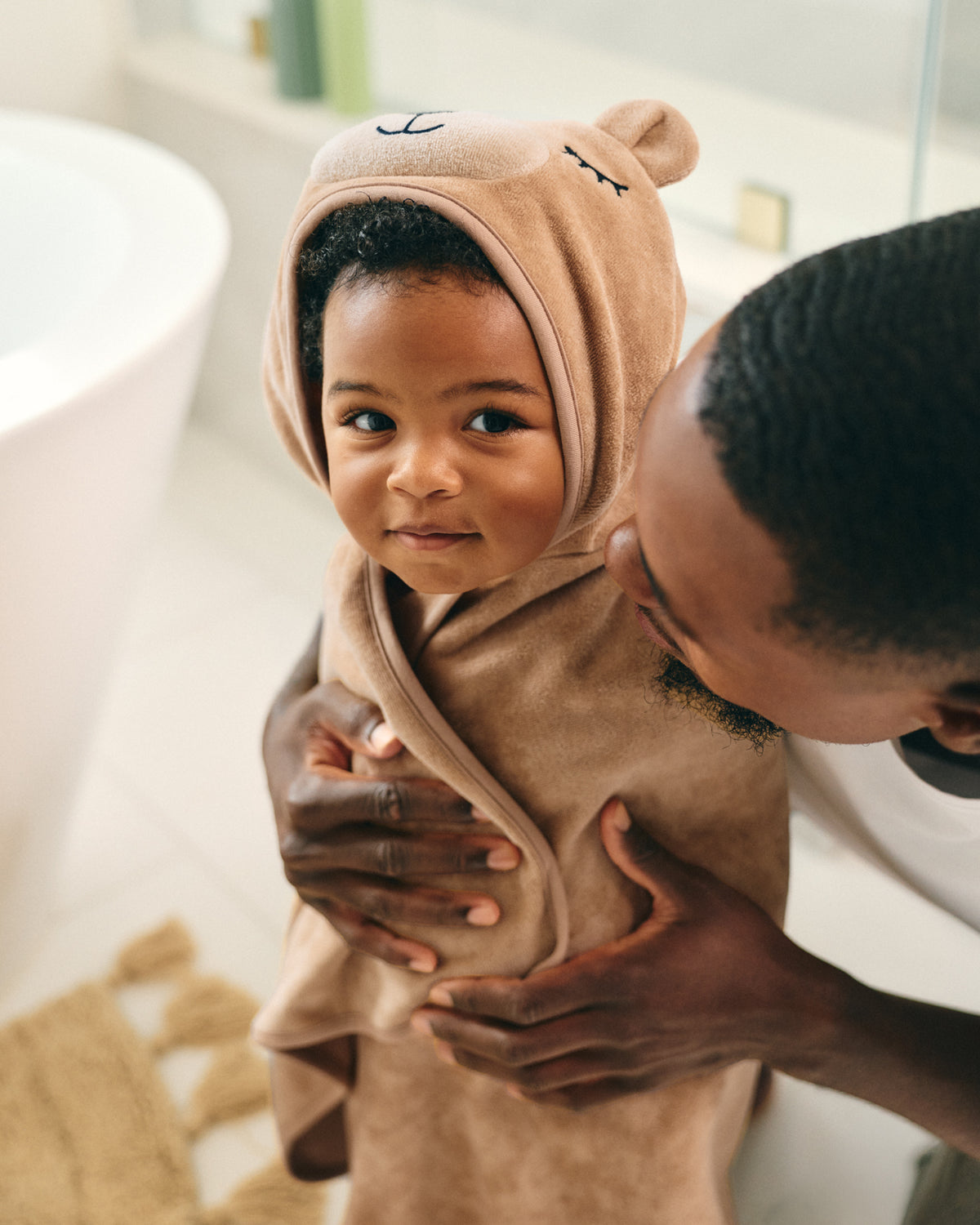 A baby wrapped in a beige plush hooded bath towel, held by an adult in a bright bathroom setting.