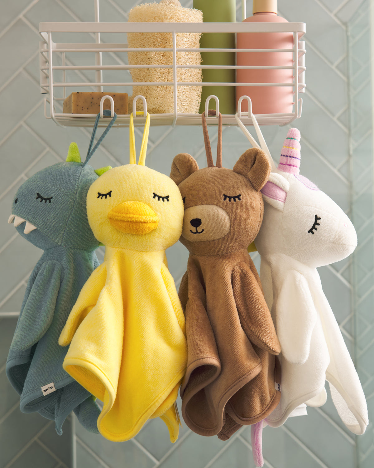 Four plush animal bath towels hanging from a white wall hook rack: a blue shark, a yellow duck, a brown bear, and a white unicorn. The towels are styled in a bathroom setting with a tiled wall and a shelf holding bath accessories in the background.