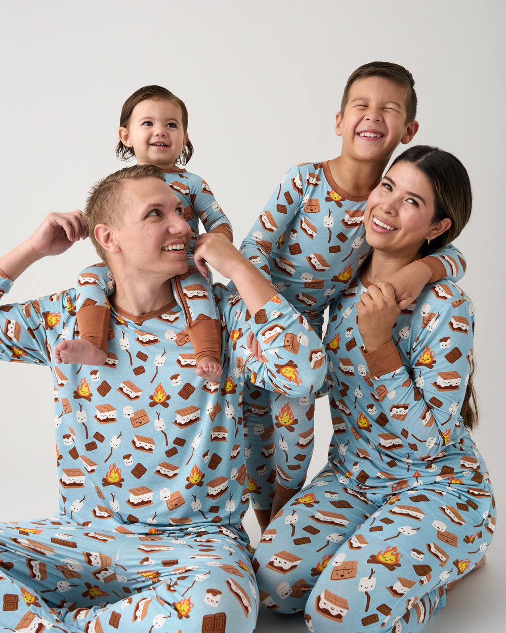 A family of four wearing matching light blue pajamas with a s'mores print, posing together against a plain light gray background.