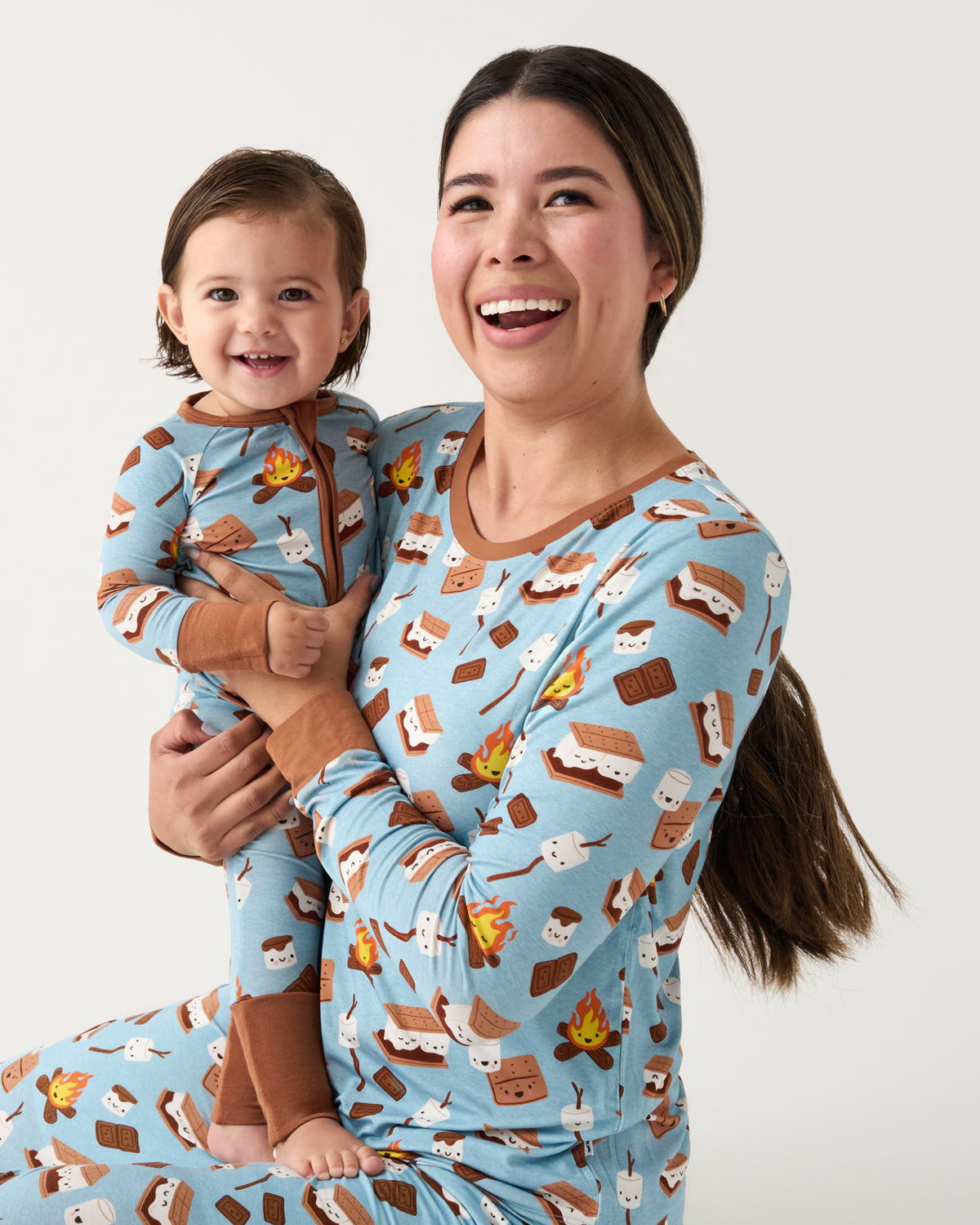 A woman and a toddler wearing matching light blue long-sleeve pajamas with a brown campfire and s'mores print, posed together against a plain light gray background.