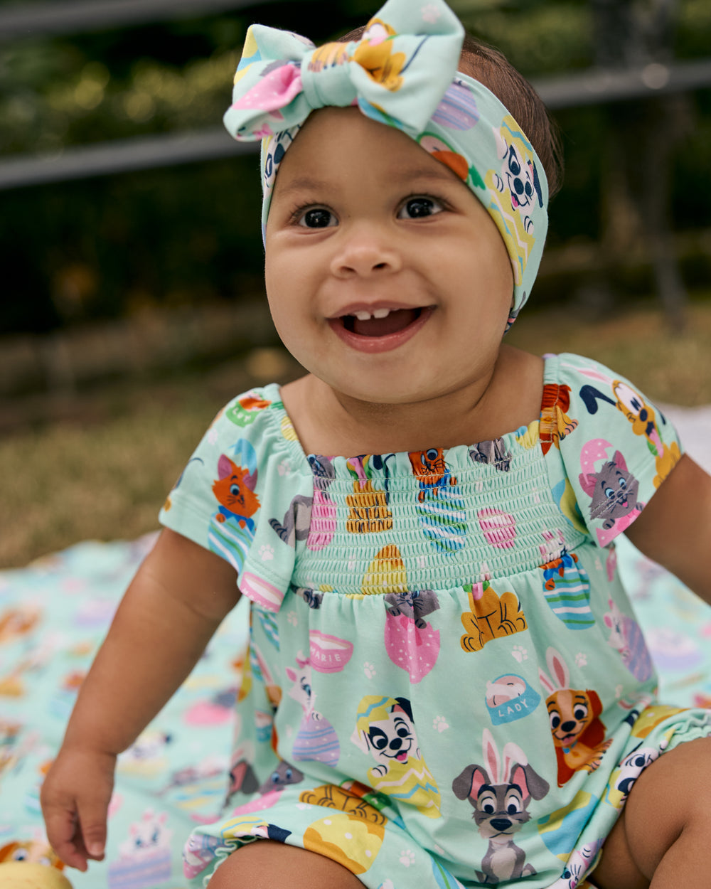 Baby wearing the Disney Peekaboo Pets Smocked Flutter Romper with greenery in the background