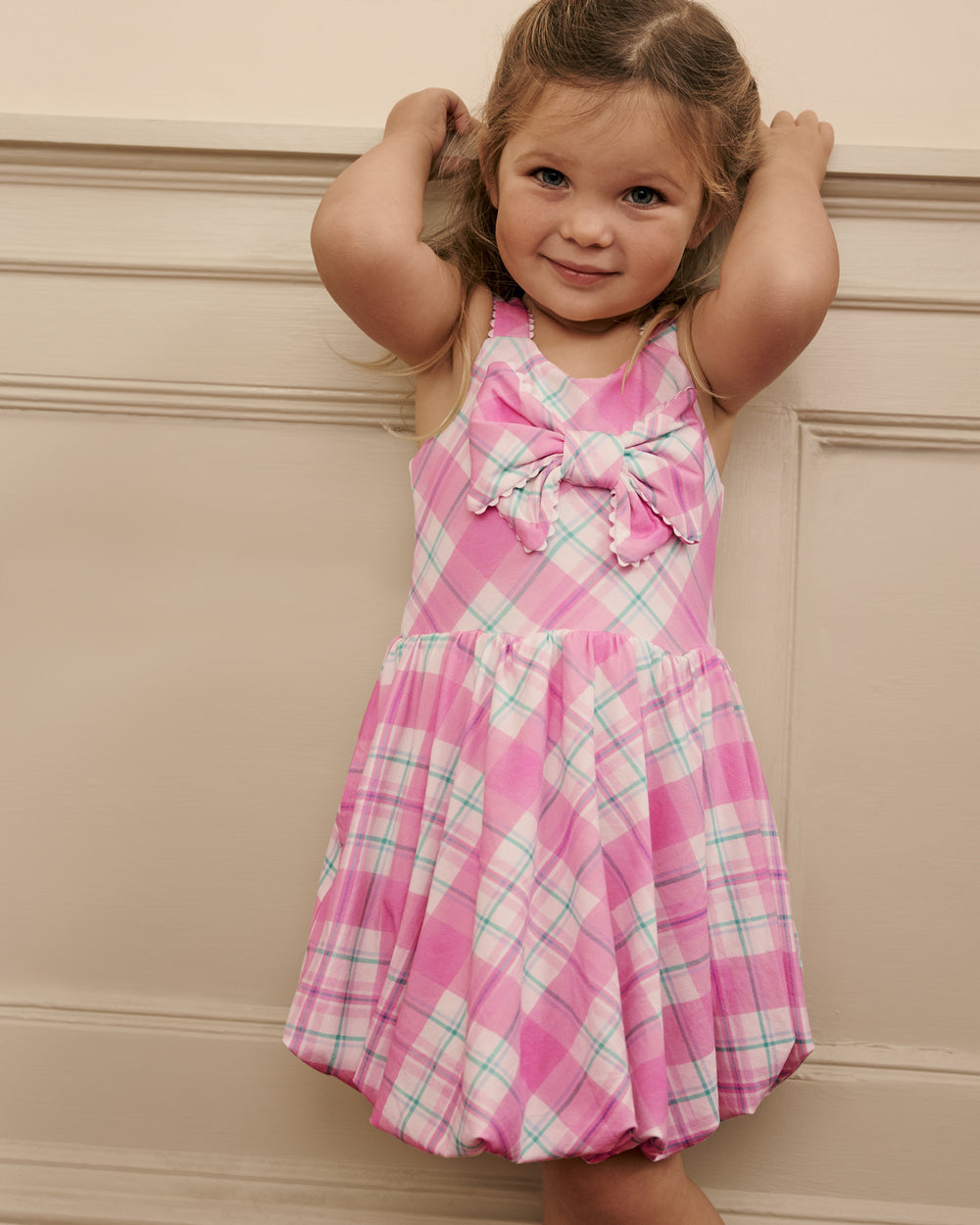 Young girl wearing a pink Springtime Plaid Bow Bubble Dress with a bow, standing against a beige wall