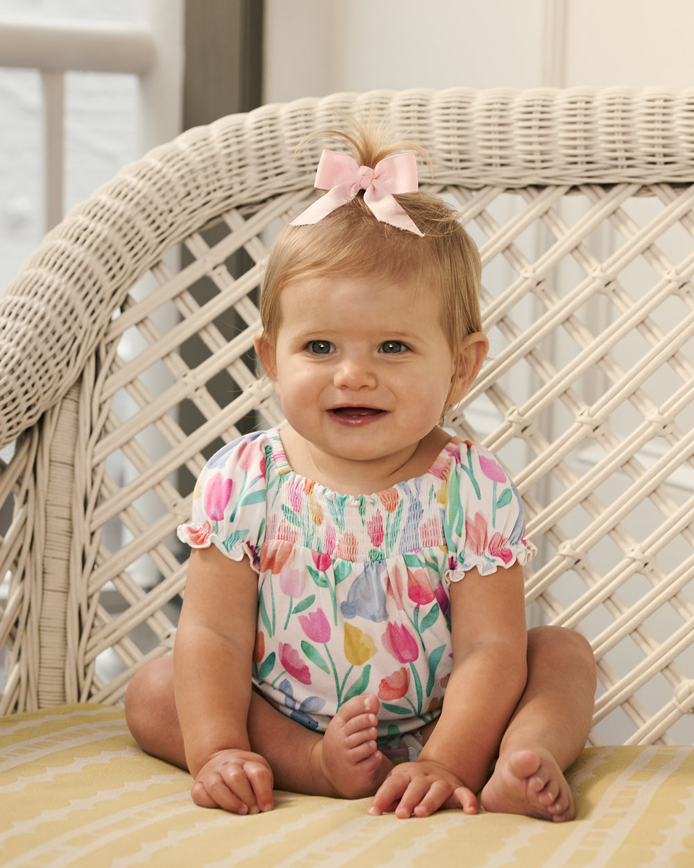 Baby girl sitting on a wicker couch wearing the Garden Hop Smocked Bubble Romper