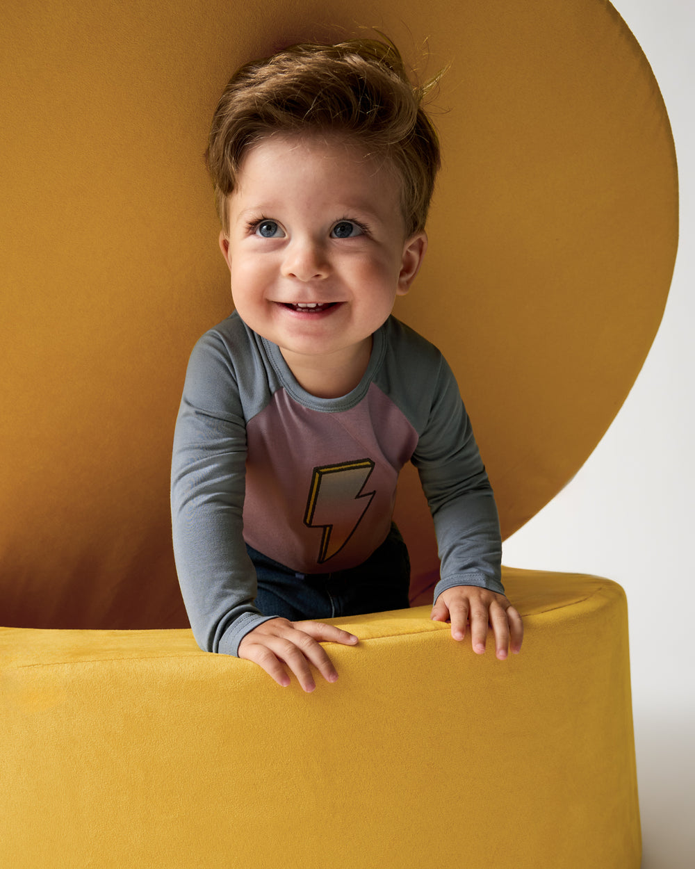 Child wearing the Bolt Raglan Tee on a yellow modular play sofa 