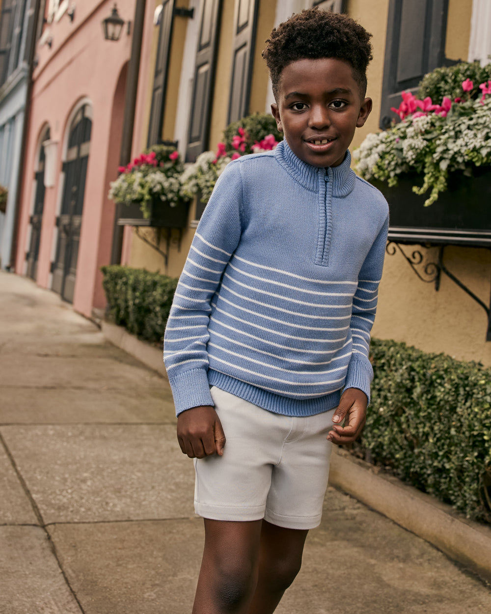 Child wearing the Infinite Blue Stripe Zip Sweater with grey boys shorts with a cityscape in the background