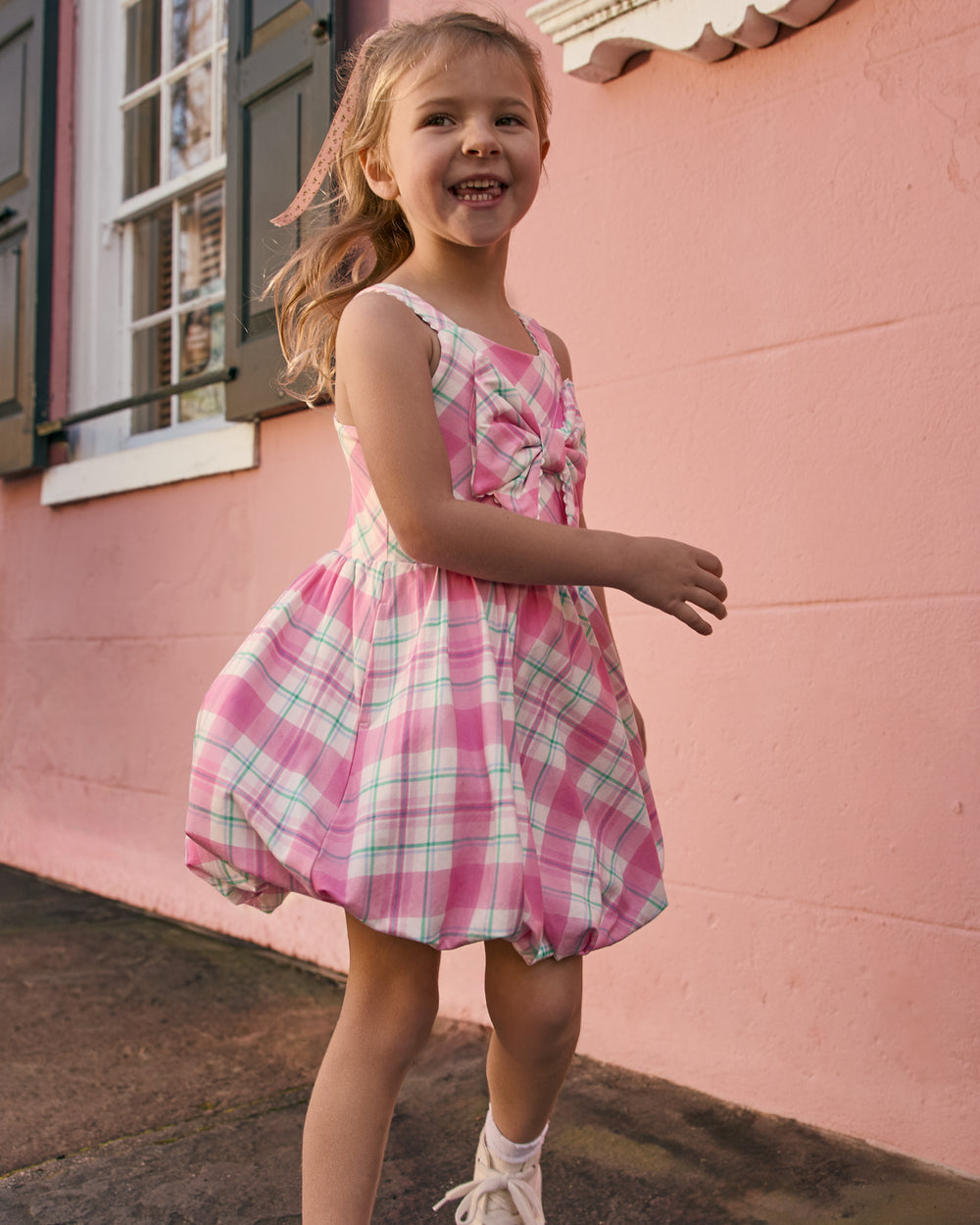 Girl wearing the Springtime Plaid Bow Bubble Dress with a pink building in the background
