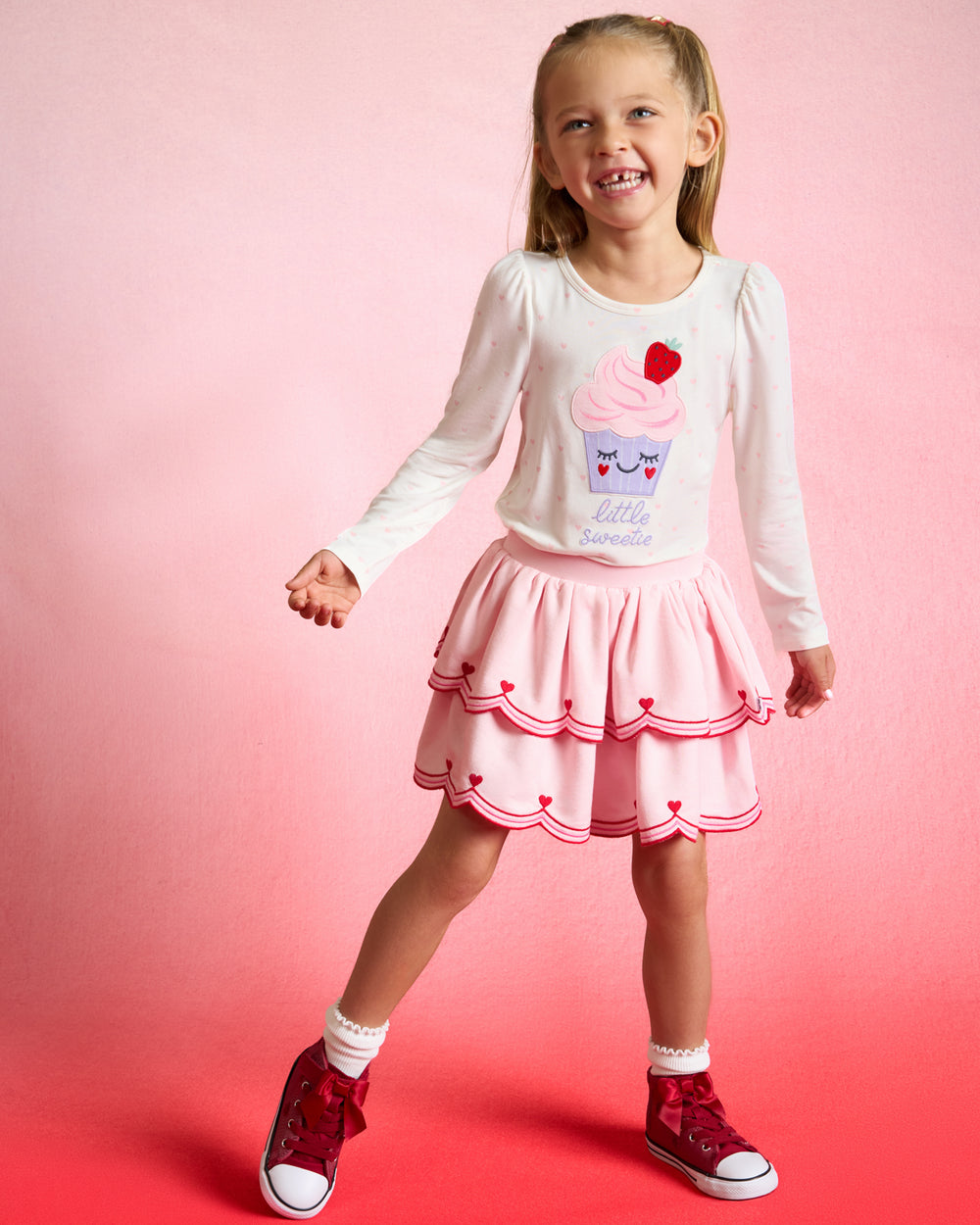 Girl wearing the Little Sweetie Puff Sleeve Tee with a pink skort on a red background