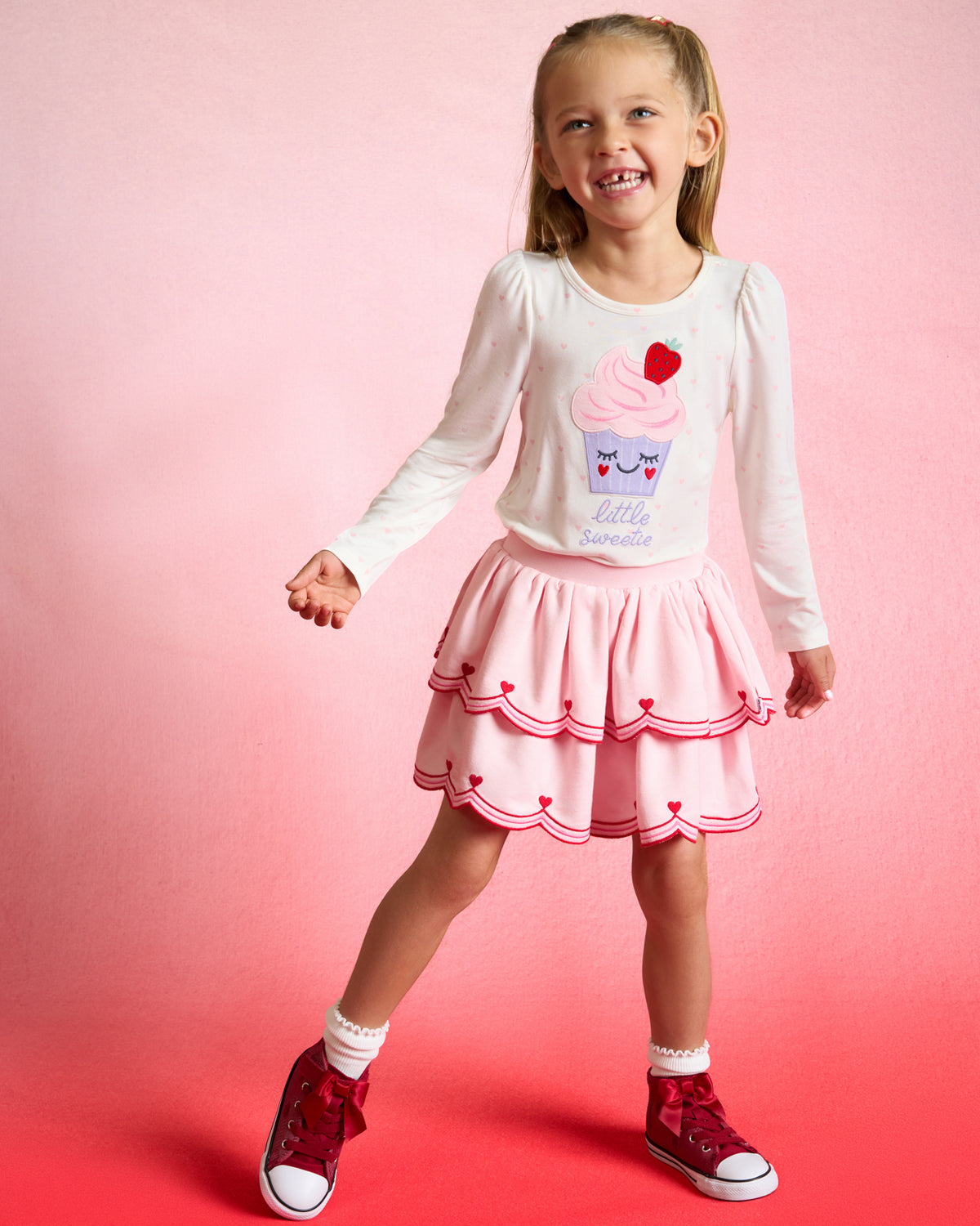 Girl wearing the Little Sweetie Puff Sleeve Tee with a pink skort on a red background