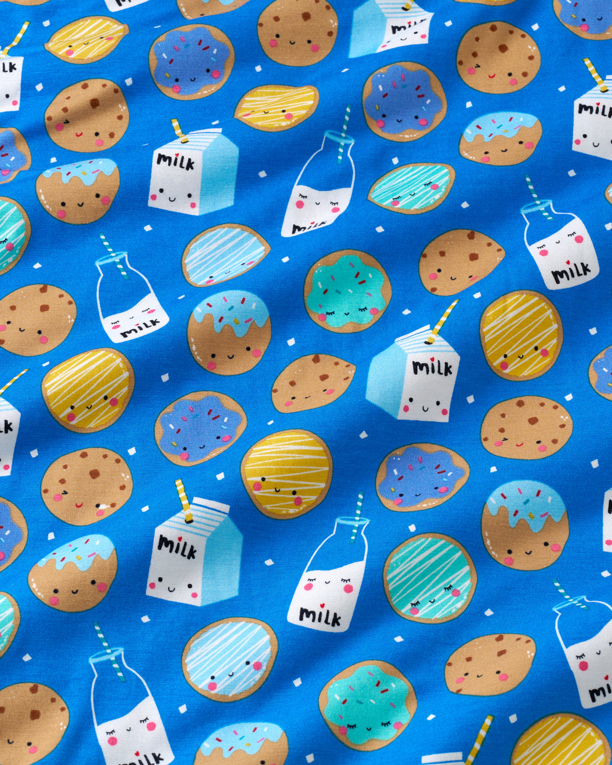 A close-up of a blue fabric featuring a repeating pattern of cute, smiling cartoon cookies, milk bottles, and milk cartons in pastel colors.