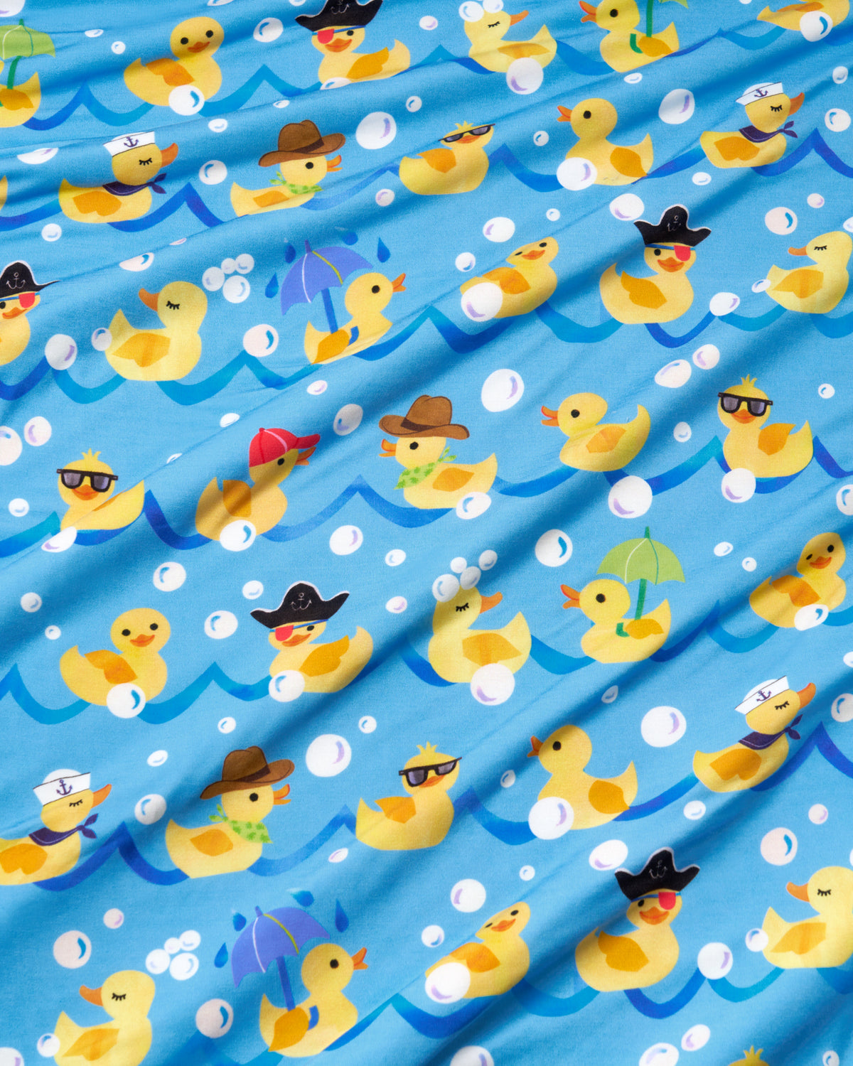 Light blue fabric with a playful pattern of yellow rubber ducks floating on wavy blue lines, decorated with white bubbles and wearing various colorful hats, sunglasses, and umbrellas.