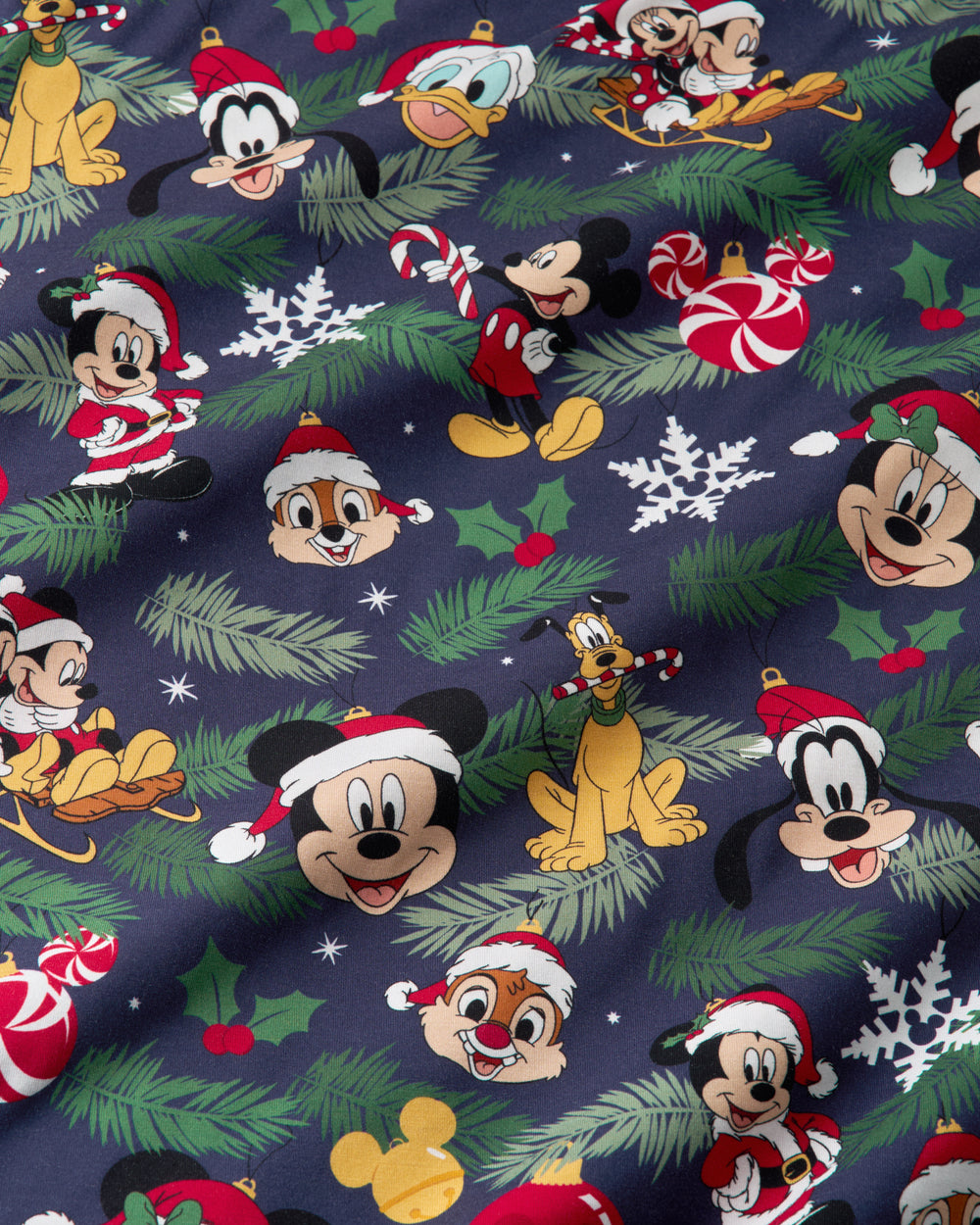 A close-up of a navy blue fabric featuring a festive Christmas print with Disney characters including Mickey Mouse, Donald Duck, Goofy, Chip 'N' Dale, Pluto, and Tweety, decorated with green pine branches, white snowflakes, red-and-white candy canes, holly leaves, and Santa hats.