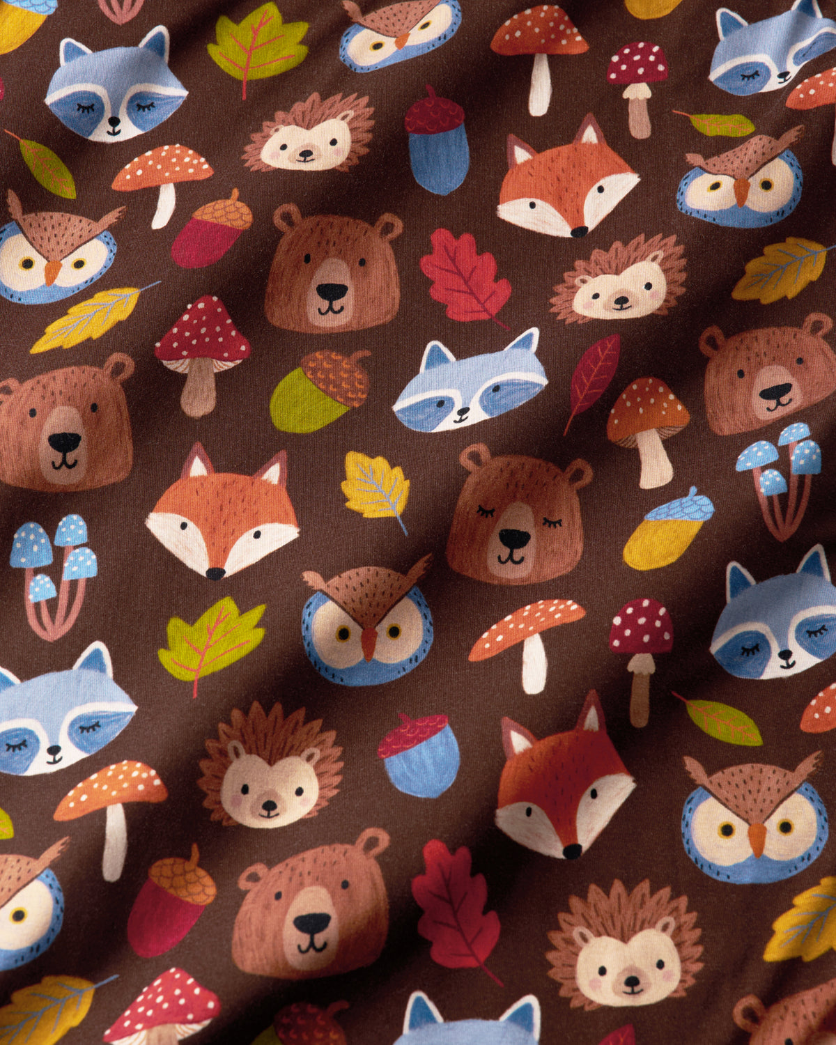 A close-up of a brown fabric with a repeating woodland pattern featuring cartoon bears, foxes, hedgehogs, owls, raccoons, mushrooms, acorns, and leaves in autumn colors.