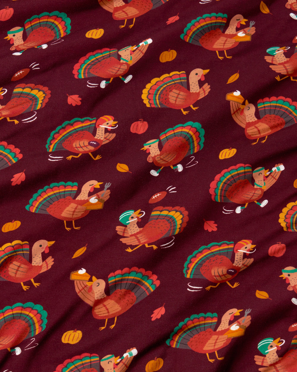 A close-up of a fabric with a repeating Thanksgiving turkey pattern on a deep burgundy background. The fabric features stylized turkeys in orange, brown, and green, along with small pumpkins and autumn leaves.