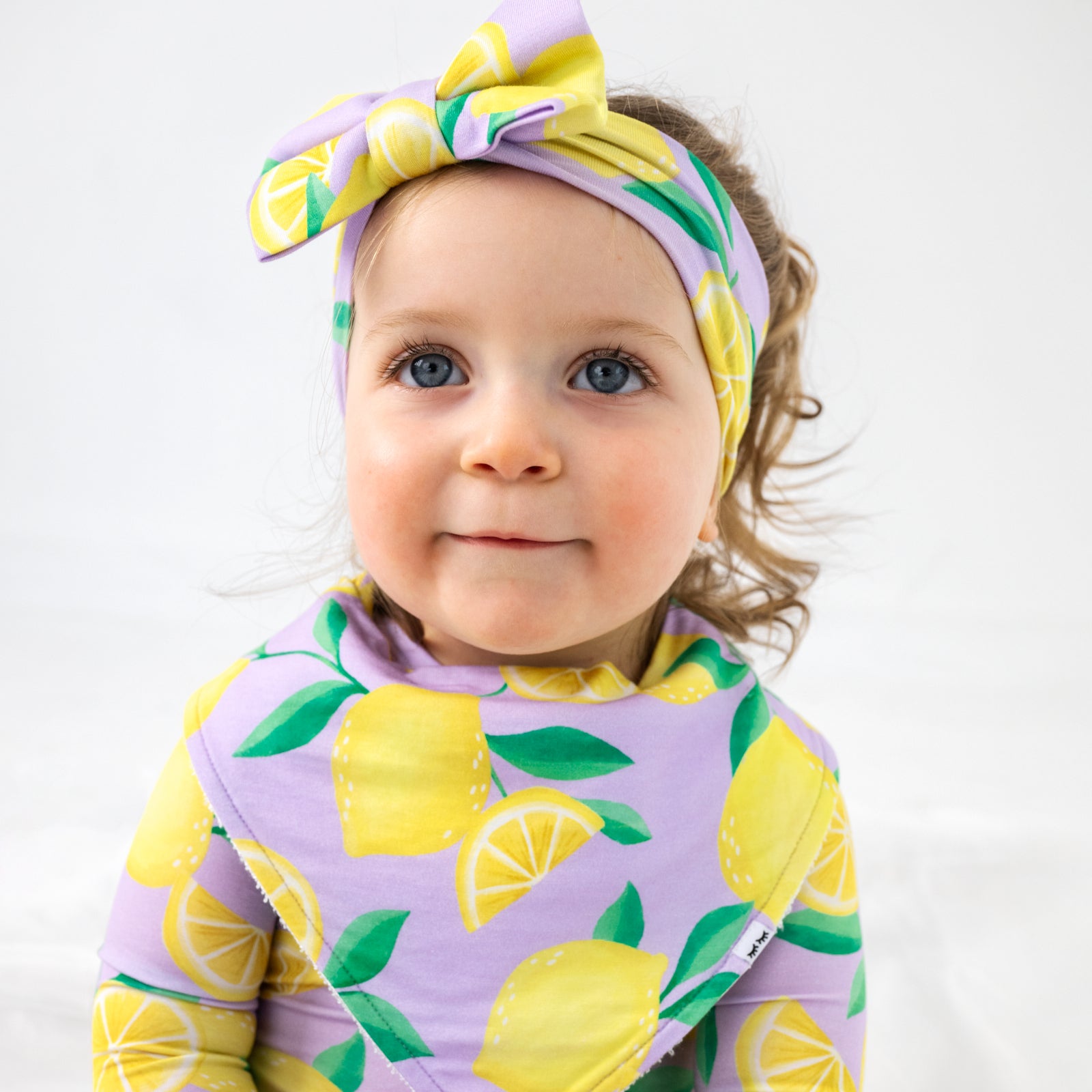 Child wearing the Lavender Sunny Citrus Bandana Bib and matching lixe bow headband