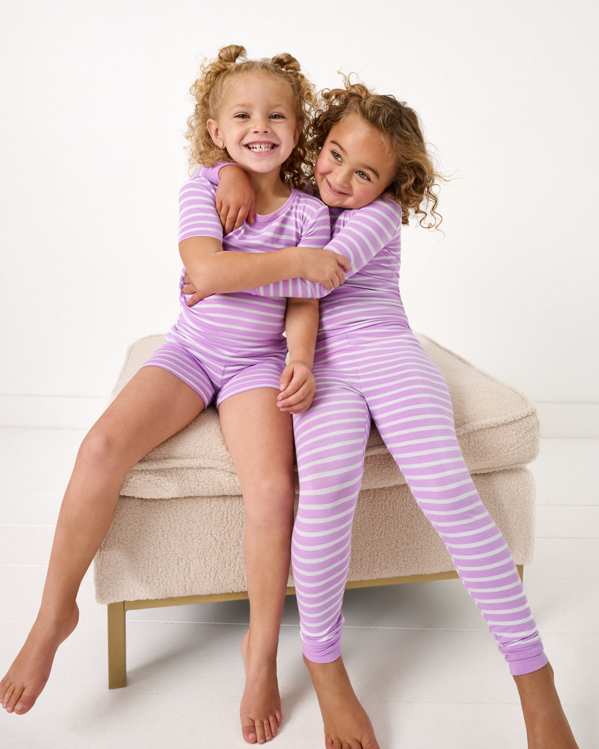 Two girls sitting on an ottoman wearing matching Sweet Orchid Stripe Two-Piece Pajama Sets