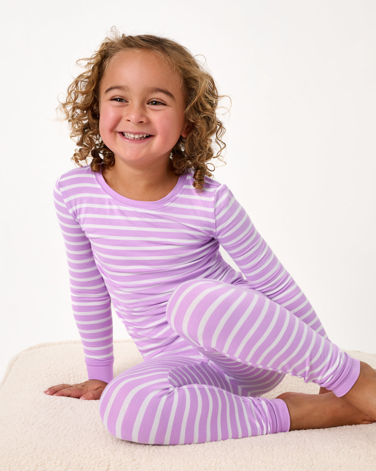 Girl sitting on a boucle ottoman wearing the Sweet Orchid Stripe Two-Piece Pajama Set