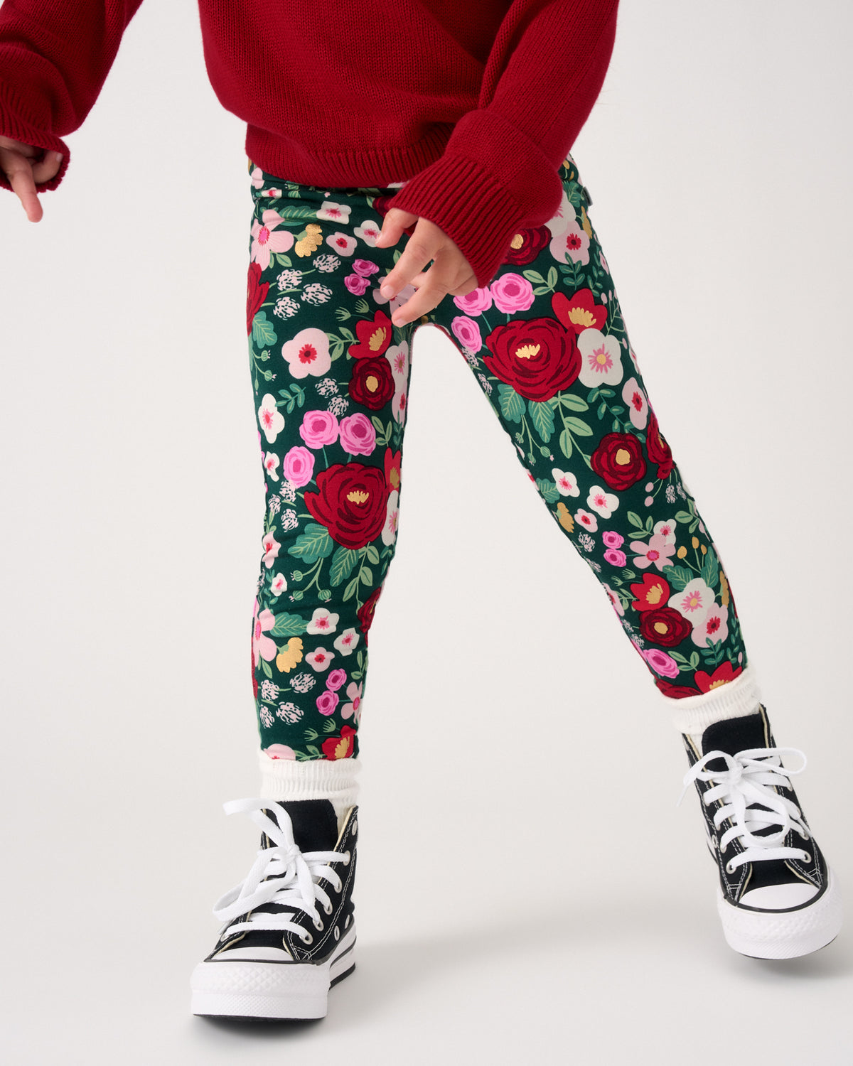 A child wearing dark green floral leggings with red, pink, and white flowers, styled with a red sweater and black high-top sneakers against a light gray studio background.