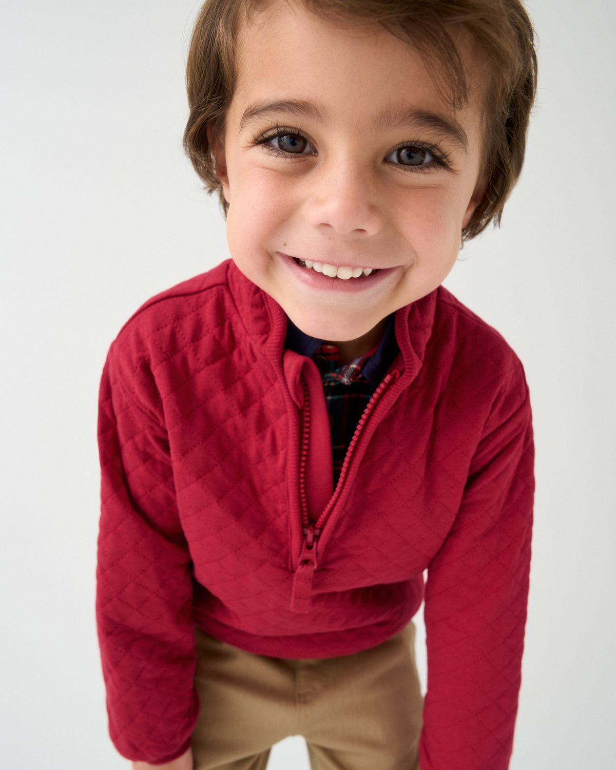A smiling young boy wearing a red quilted half-zip pullover sweater, tan pants, and a patterned shirt collar, photographed against a clean light gray studio background.