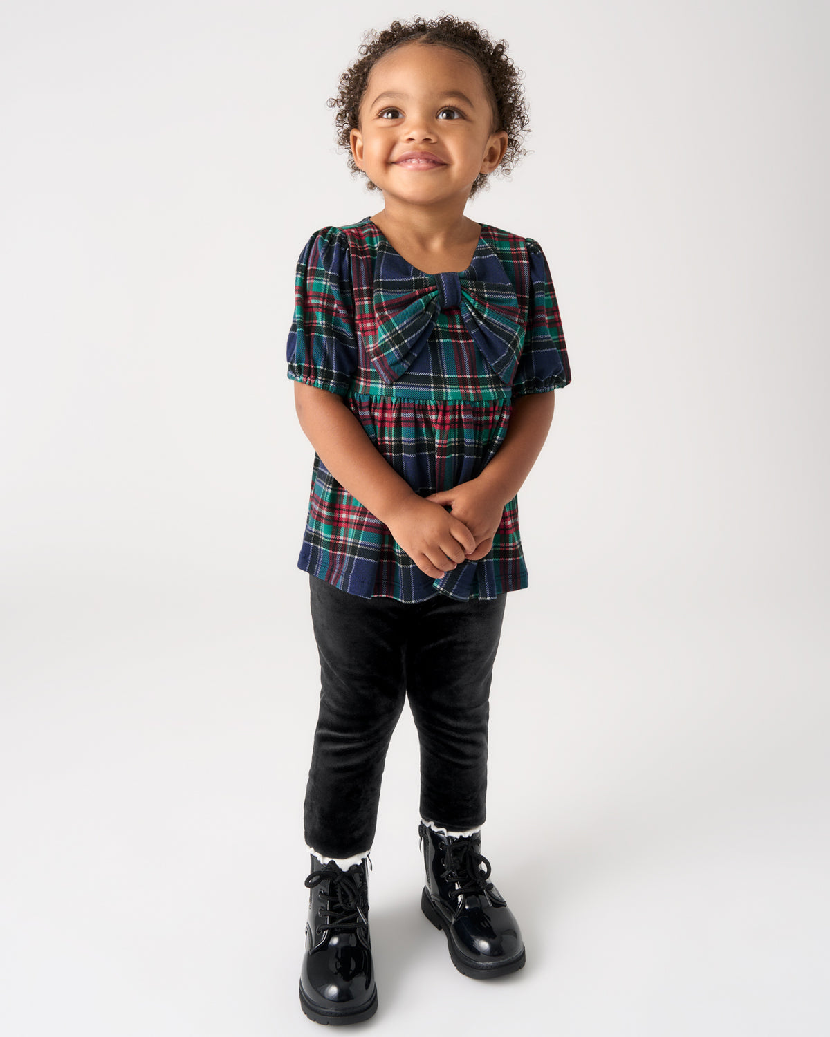A toddler girl wearing a short-sleeve plaid top with a large bow at the collar, paired with black pants and black boots, standing against a light gray studio background.