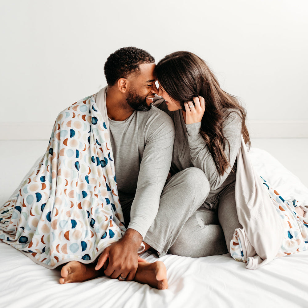 Click to see full screen - Man and woman cuddling under a Luna Neutral oversized cloud blanket. They are wearing Heather Gray women's and men's pajama tops and matching pj pants