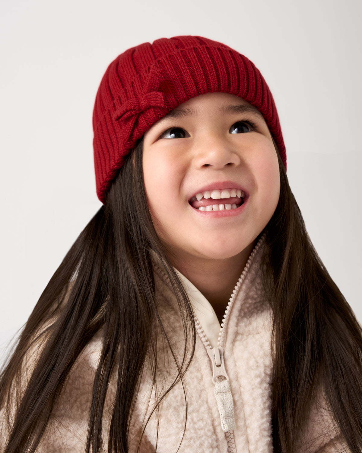 A young girl with long dark hair smiling and looking upward, wearing a red ribbed knit beanie with a bow detail and a cream fleece zip-up jacket, against a plain light gray background.