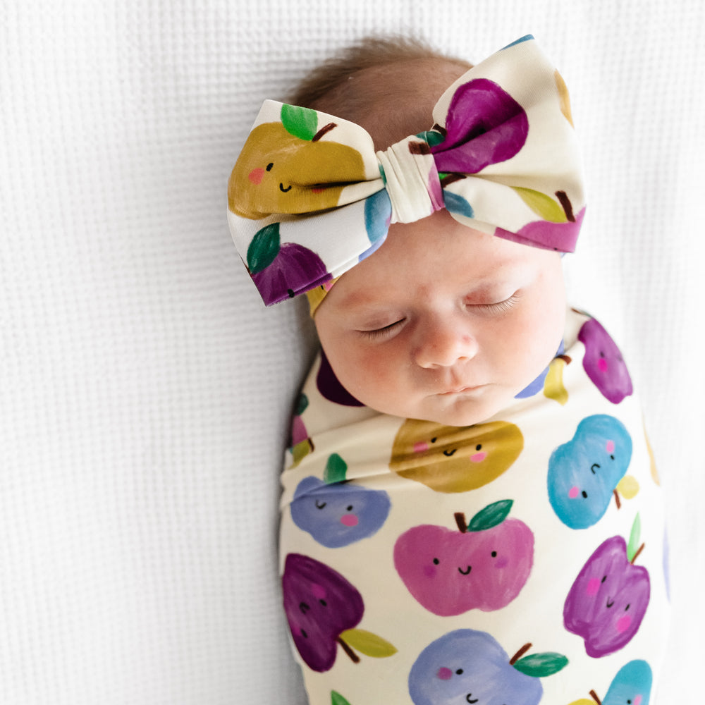 Close up image of a child laying on a bed swaddled in a Berry Apple of my Eye swaddle and luxe bow headband set