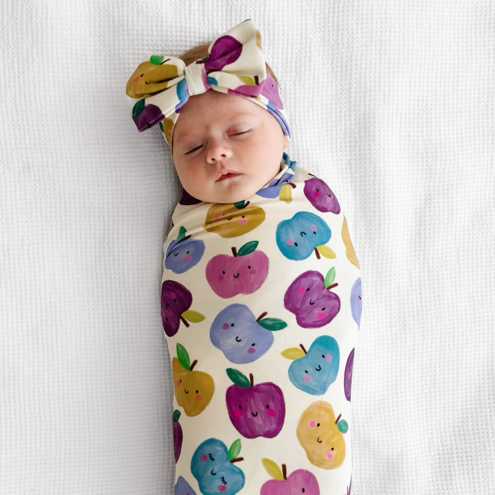 Alternate image of a child laying on a bed swaddled in a Berry Apple of my Eye swaddle and luxe bow headband set