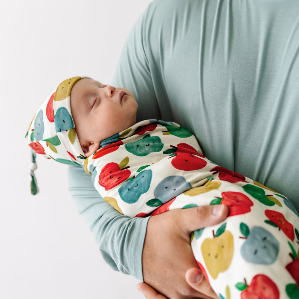 Click to see full screen - Man holding a child swaddled in a Red Apple of My Eye swaddle and hat set