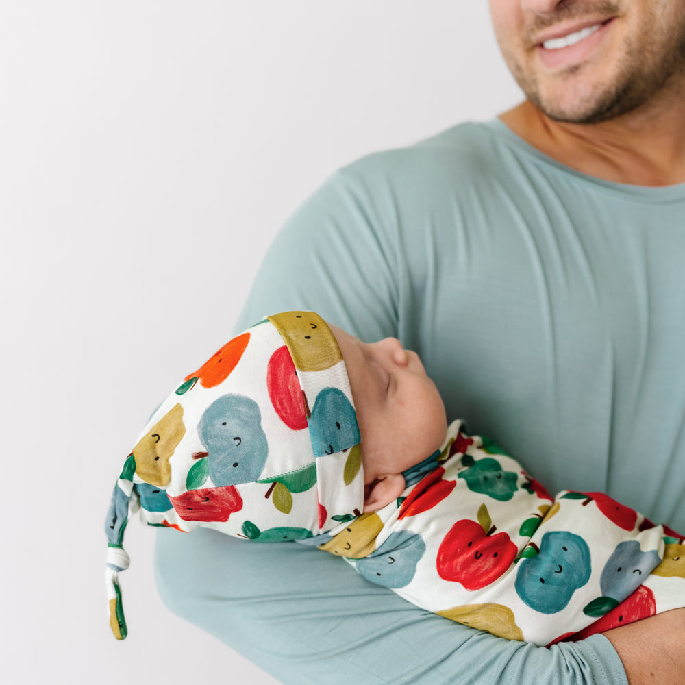 Alternate image of a man holding a child swaddled in a Red Apple of My Eye swaddle and hat set