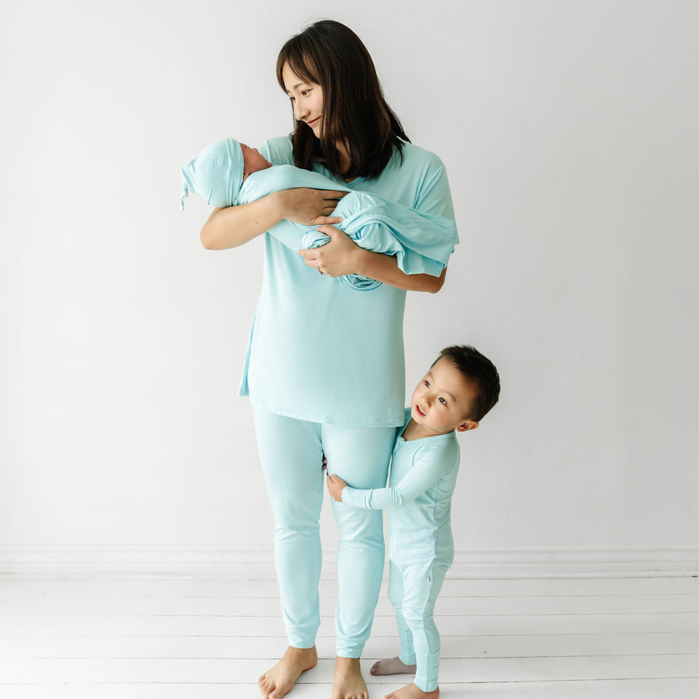 Mom posing with her two children. The baby in her arms is wearing an Aquamarine swaddle and hat set and her son holding on to her legs is wearing an Aquamarine zippy. Mom is wearing an Aquamarine women's short sleeve pajama top paired with women's Aquamarine pajama bottoms