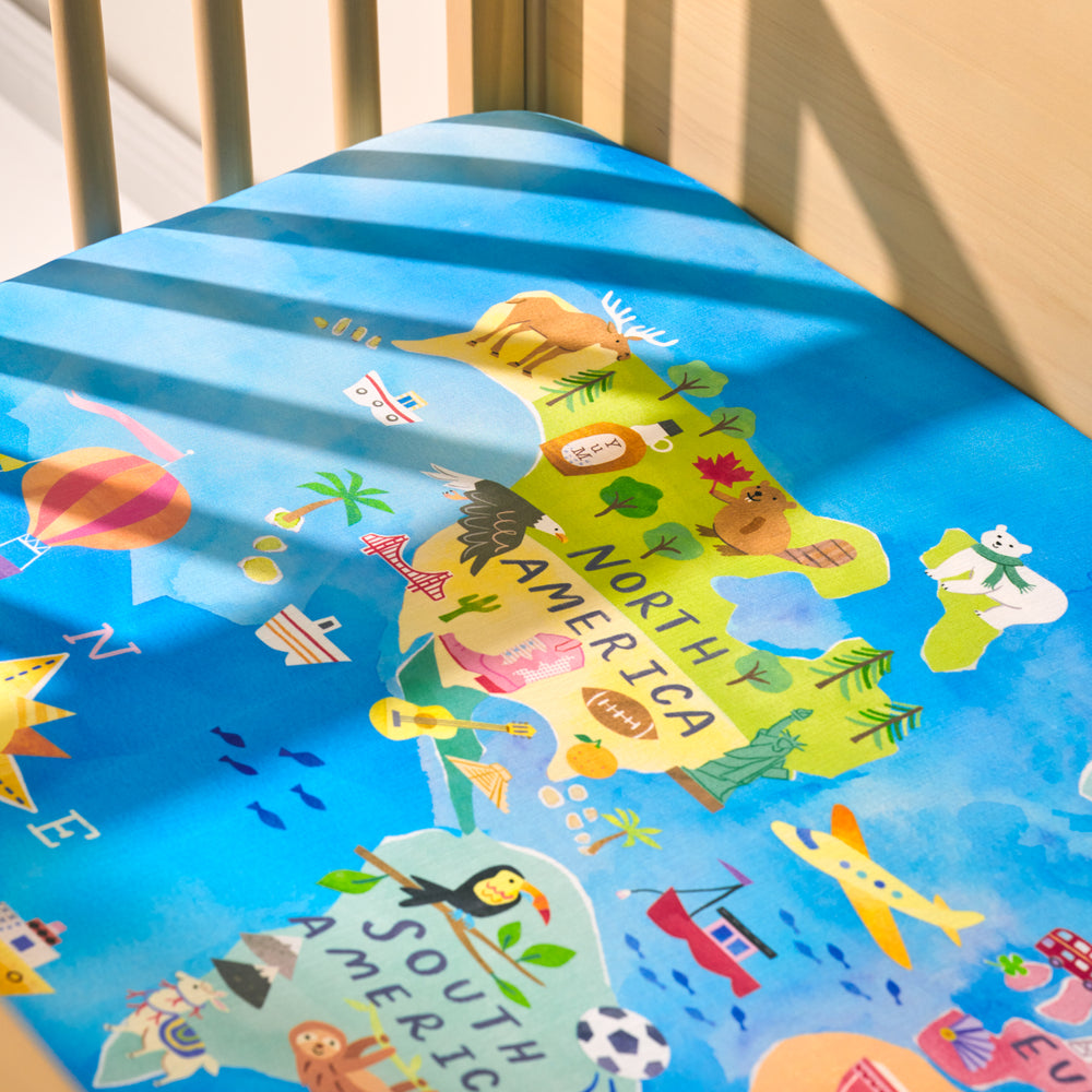 Close up view of an Around the World Fitted Crib Sheet on a mattress in a wooden crib detailing the North and South America illustrations