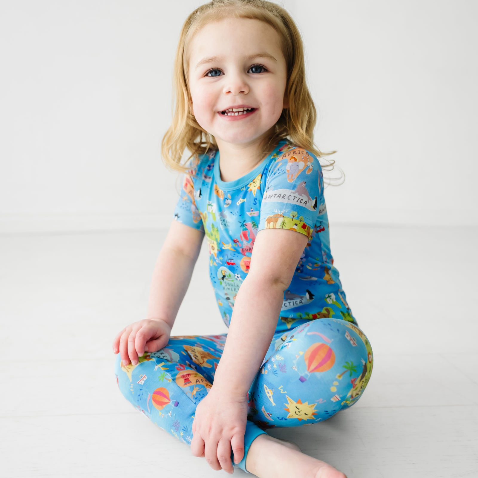 Smiling child sitting on the ground wearing an Around the World Two-Piece Short Sleeve Pajama Set