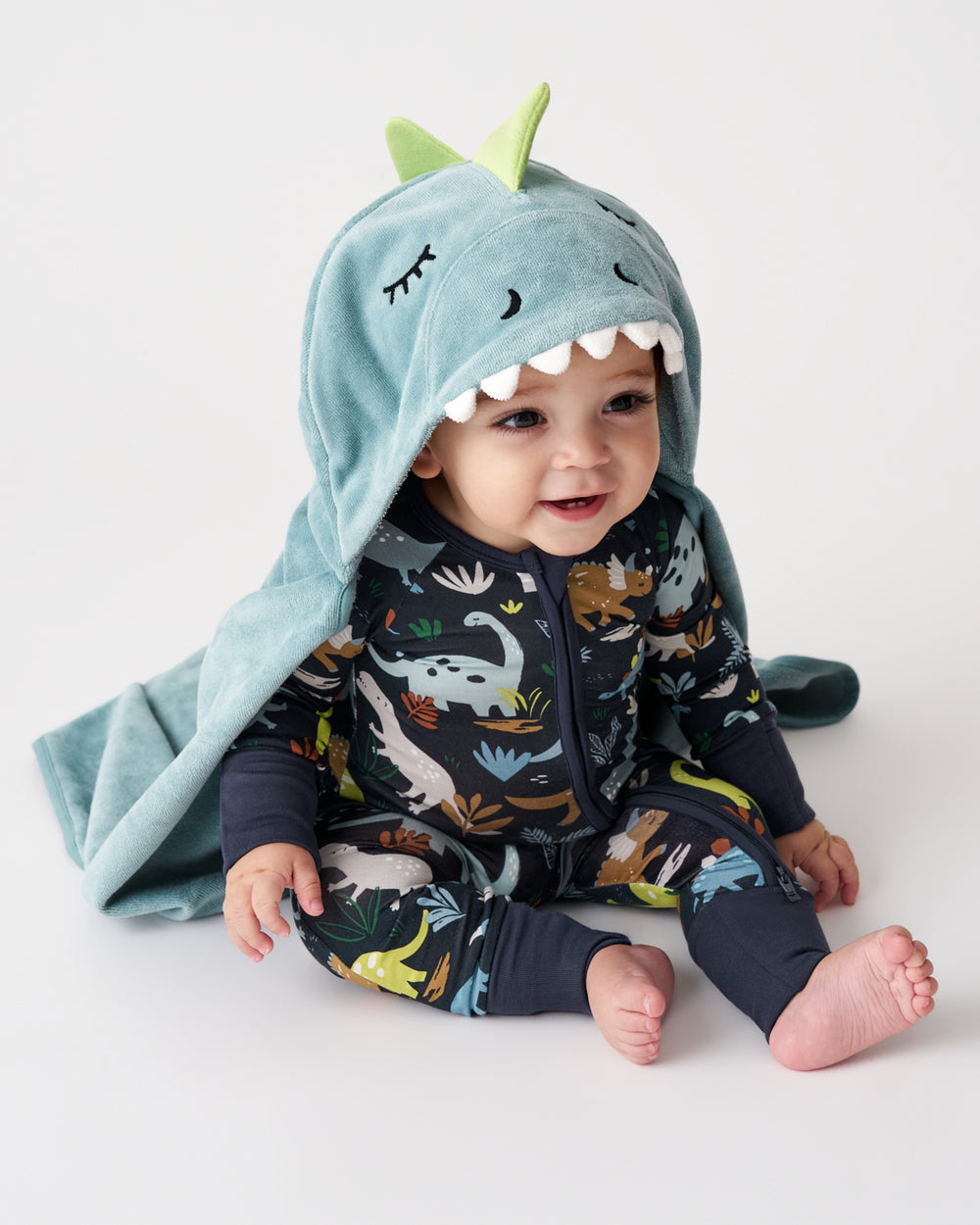 A baby wearing a dark navy dinosaur-print zip-up one-piece and a light blue hooded towel with green dinosaur ears and white teeth, sitting against a plain white studio background.