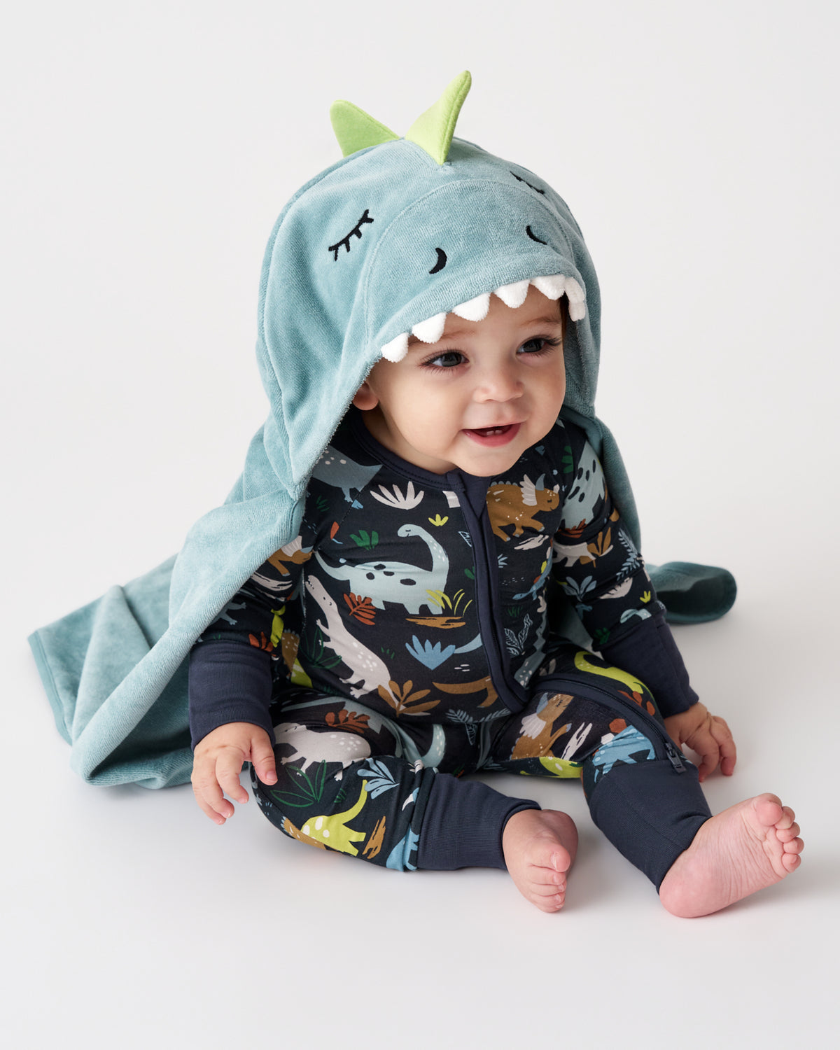 A baby wearing a dark navy dinosaur-print zip-up one-piece and a light blue hooded towel with green dinosaur ears and white teeth, sitting against a plain white studio background.