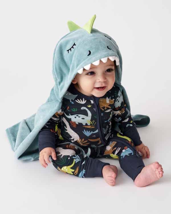 A baby wearing a dark navy dinosaur-print zip-up one-piece and a light blue hooded towel with green dinosaur ears and white teeth, sitting against a plain white studio background.