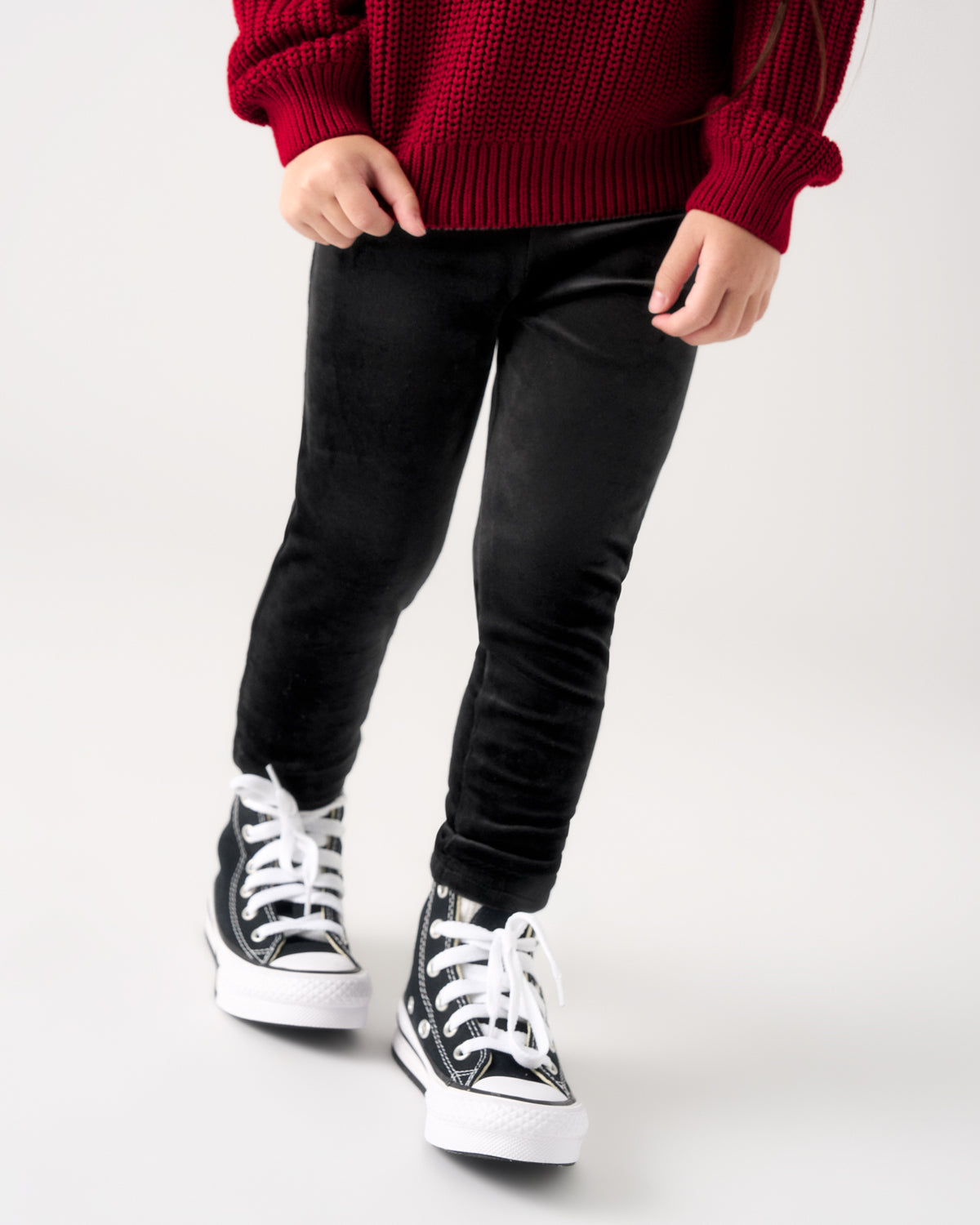 A child wearing black velvet pants, a red knit sweater, and black and white canvas sneakers, shown from the waist down against a light gray studio background.