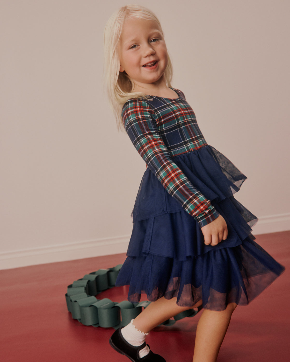 A young blonde girl wearing a navy blue tiered tulle dress with a red, green, and blue plaid long-sleeve top, standing on a red floor in front of a beige wall.