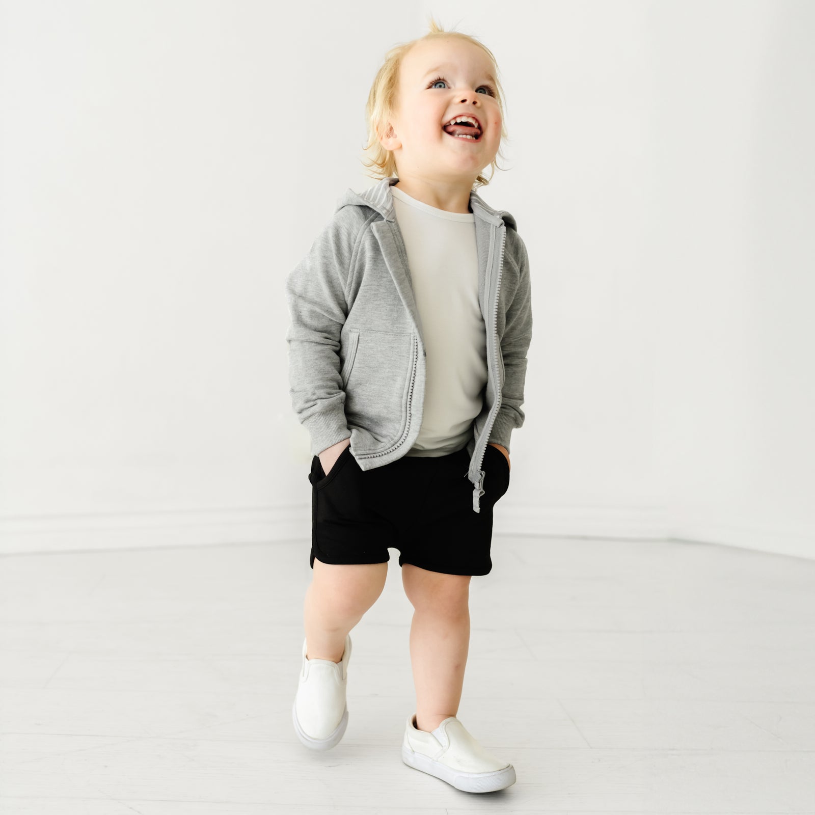 Child looking up and laughing, wearing Black Dolphin Shorts and coordinating white boys shirt and light heather gray boys zip hoodie