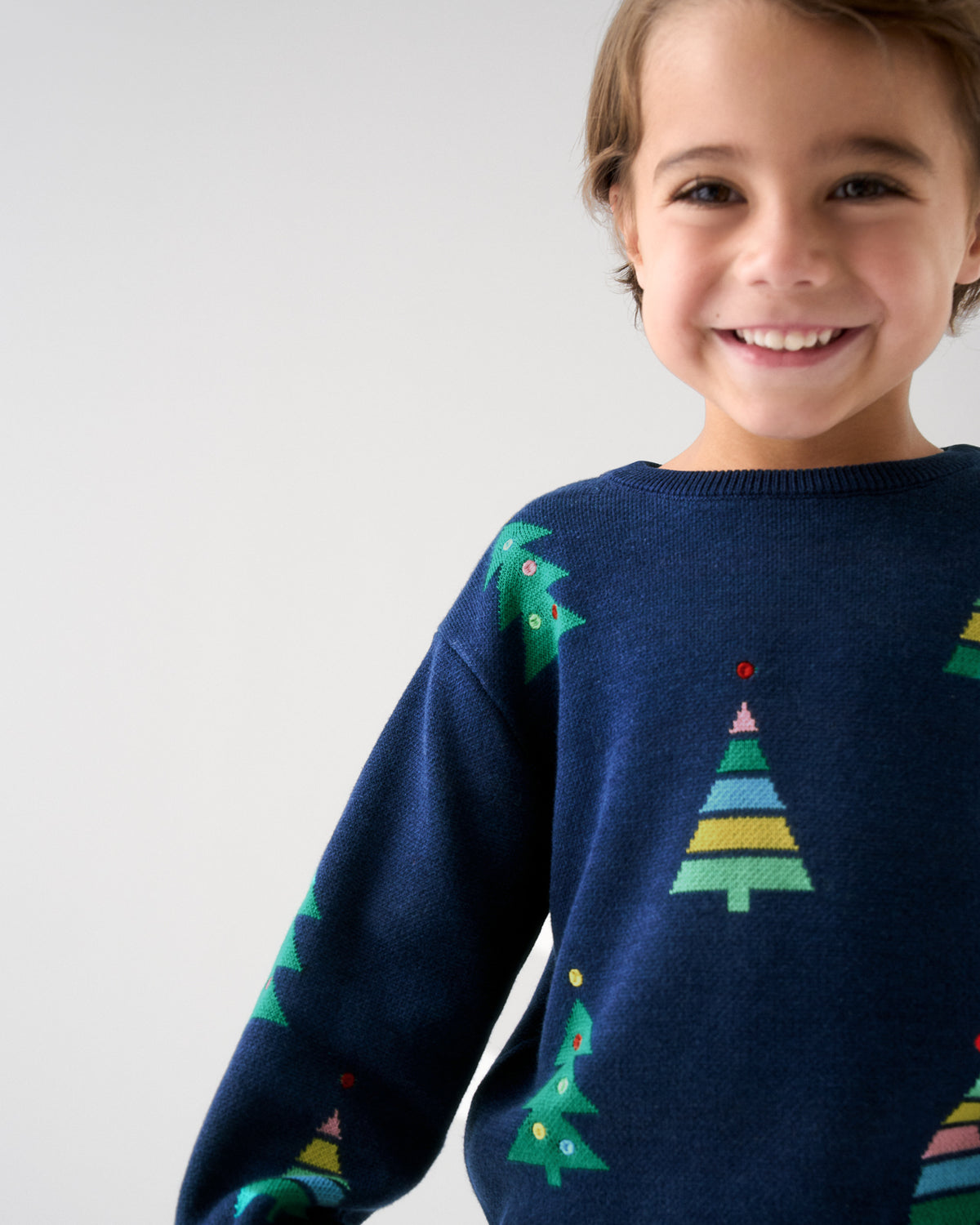 A smiling young boy wearing a navy blue knit sweater with colorful Christmas tree patterns, photographed against a clean light gray background.