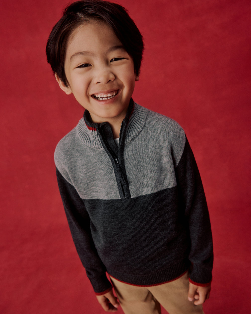 A smiling young boy wearing a color-blocked quarter-zip sweater, standing against a solid red studio backdrop.