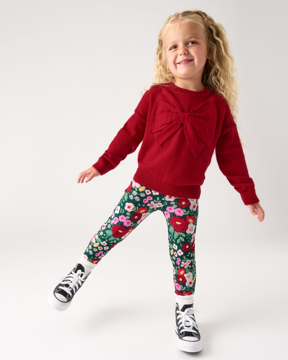 A young girl with blonde curly hair is wearing a red knit sweater with a large front bow, green floral leggings, and black and white sneakers, posing against a light gray studio background.
