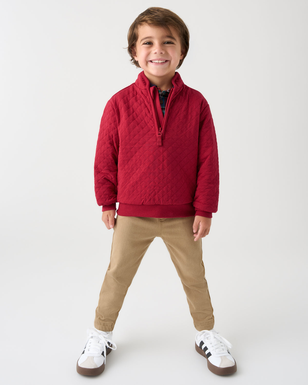 A young boy smiling and posing in a studio setting, wearing a red quilted half-zip pullover sweater, tan pants, and white sneakers with black stripes.
