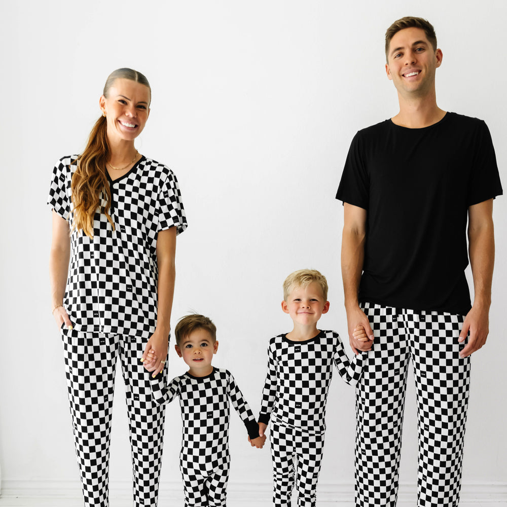 Click to see full screen - Family of four holding hands wearing matching Cool Checks printed pajamas. Mom is wearing women's Cool Checks printed women's pajama top and matching women's pajama bottoms. Dad is wearing Cool Checks printed men's pajama bottoms paired with a solid black short sleeve pajama top. Children are matching wearing Cool Checks printed two piece pajama set and crescent zippy.