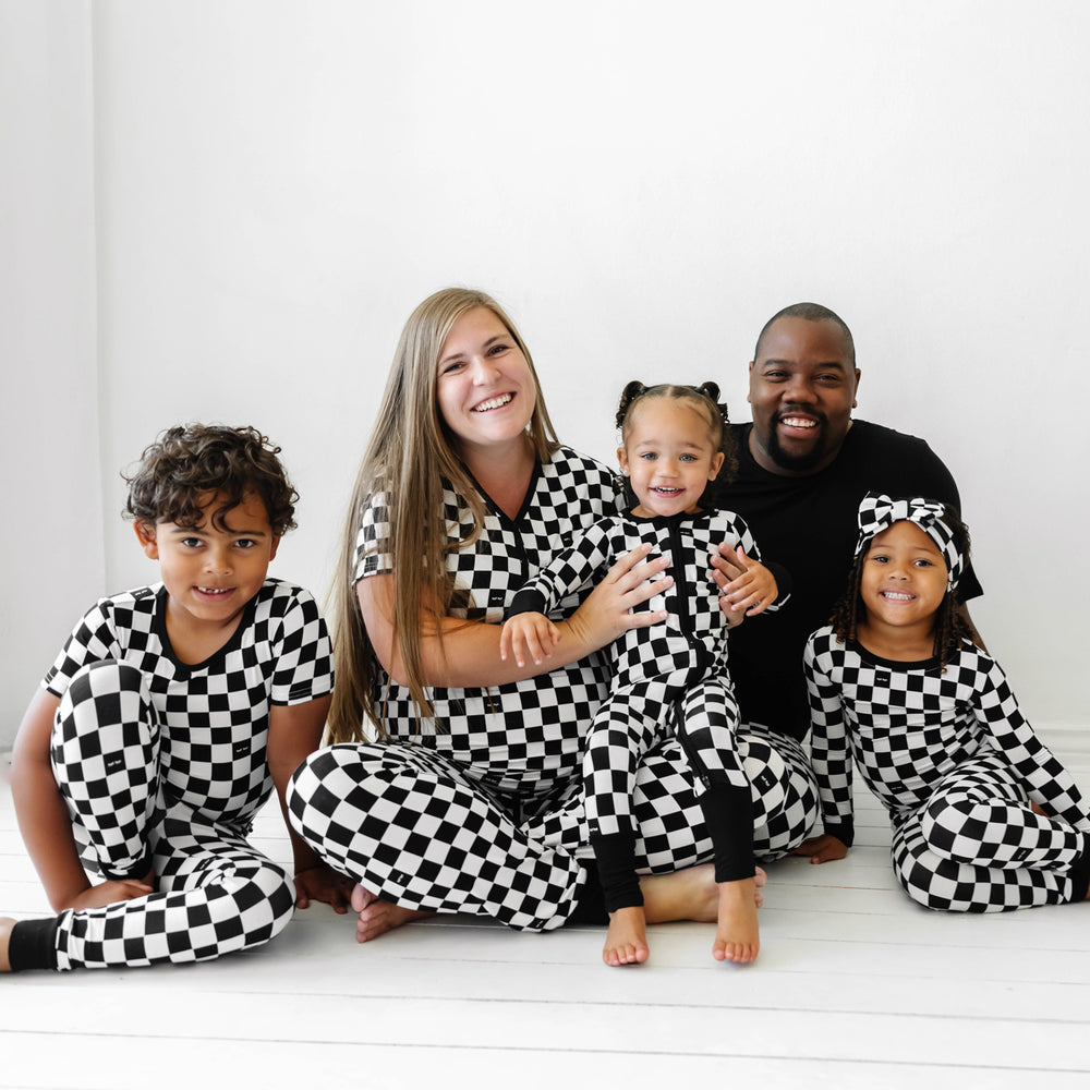Family of five sitting together wearing matching Cool Check printed pajama sets. Mom is wearing women's Cool Checks printed women's pajama top and matching women's pajama bottoms. Dad is wearing Cool Checks printed men's pajama bottoms paired with a solid black short sleeve pajama top. Children are matching wearing Cool Checks printed two piece pajama set paired with a luxe bow headband, two piece short sleeve pajama set, and crescent zippy.