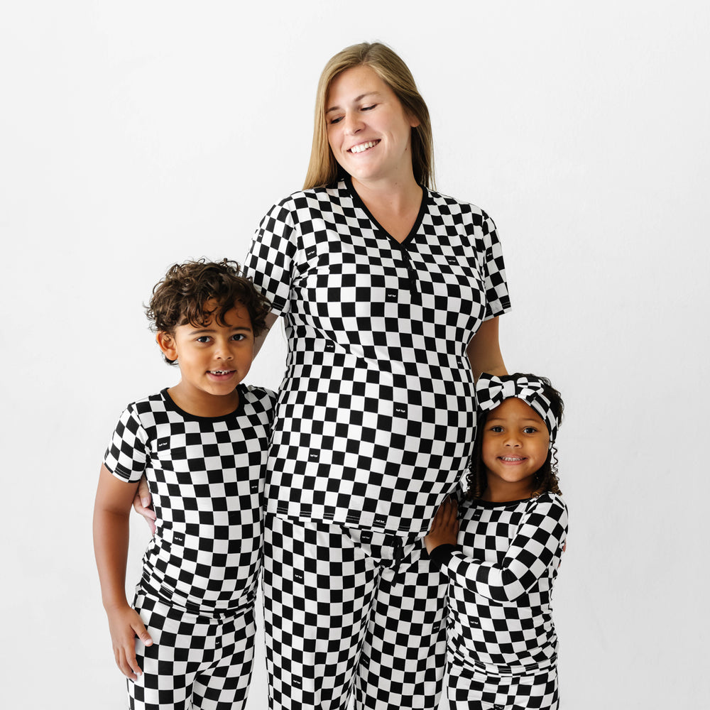 Mom posing with her two children wearing matching Cool Checks printed pajama sets. Mom is wearing Cool Checks printed women's pajama top and women's pajama pants. Kids are wearing Cool Check printed pajamas in two piece styles. Her daughter paired her Cool Checks printed pajama set with a matching luxe bow headband.