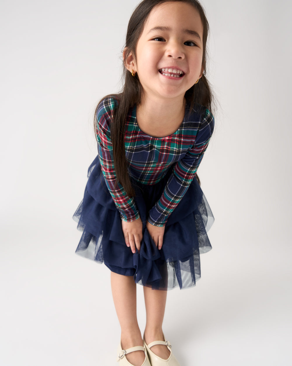 A young girl smiling and bending forward, wearing a navy blue tulle skirt dress with a long-sleeve plaid top in red, green, and blue, and cream-colored Mary Jane shoes, against a plain light gray background.