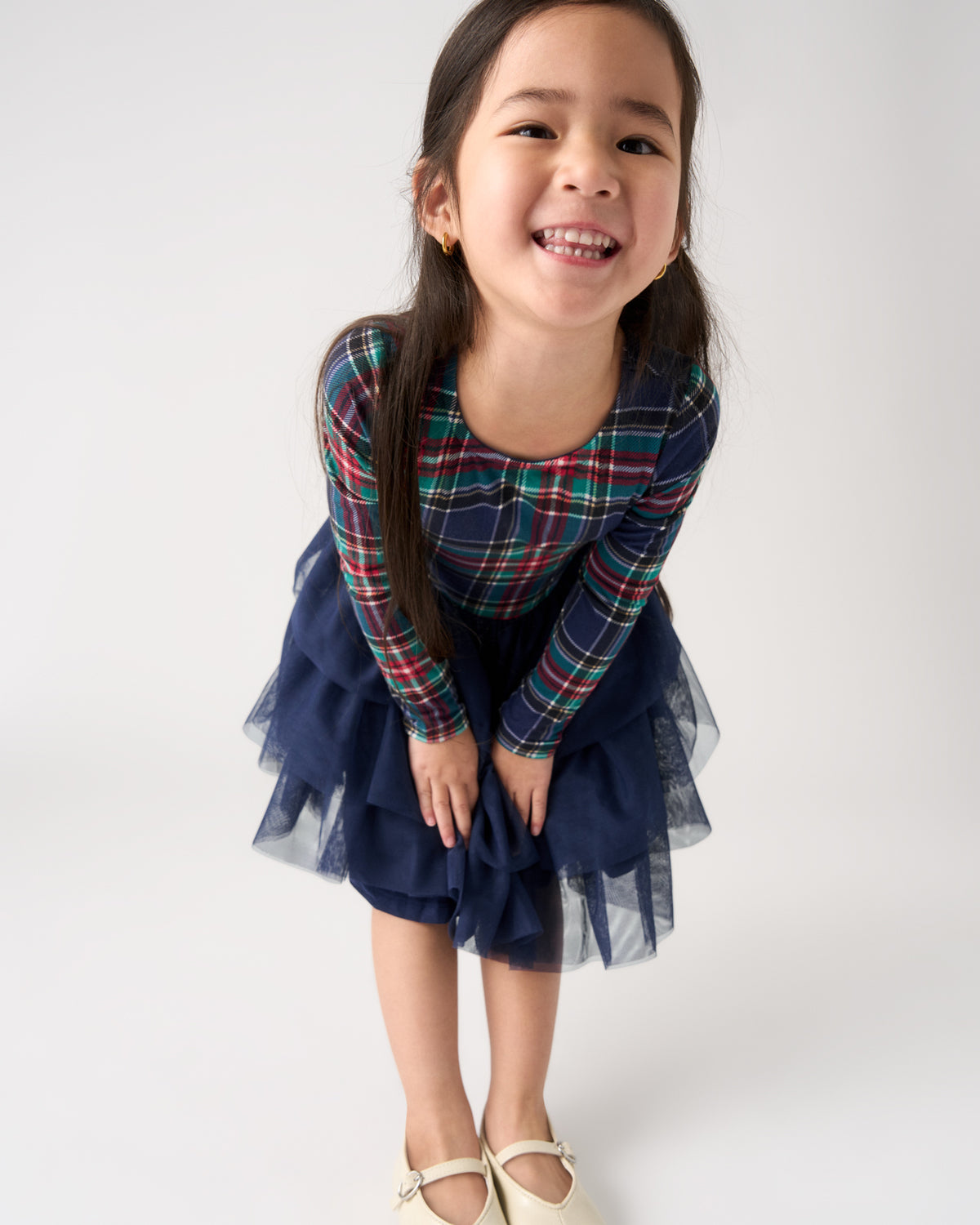 A young girl smiling and bending forward, wearing a navy blue tulle skirt dress with a long-sleeve plaid top in red, green, and blue, and cream-colored Mary Jane shoes, against a plain light gray background.