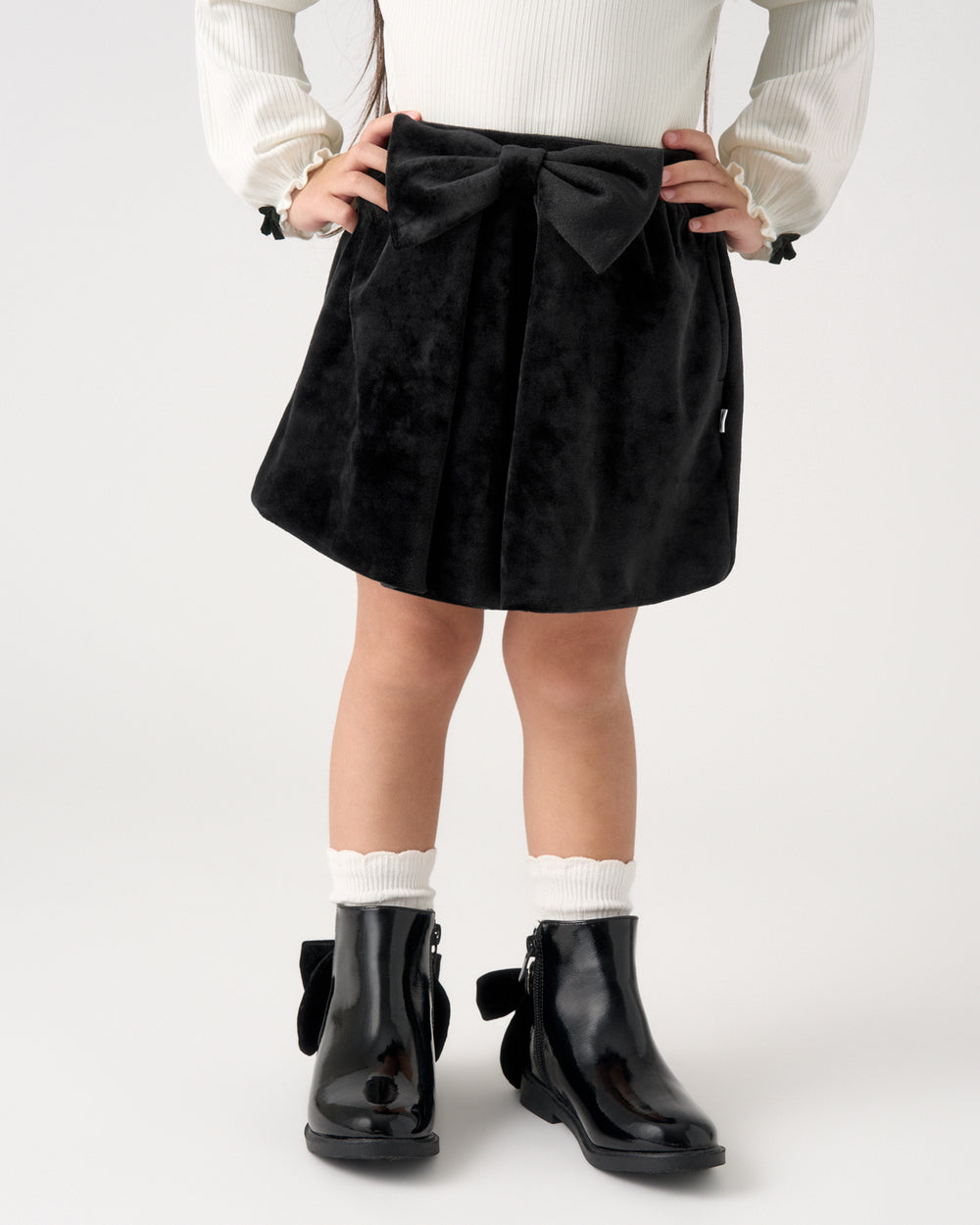 A child wearing a black velvet skirt with a large bow at the waist, paired with a white ribbed sweater, white socks, and black glossy ankle boots, shown from the waist down against a light gray studio background.